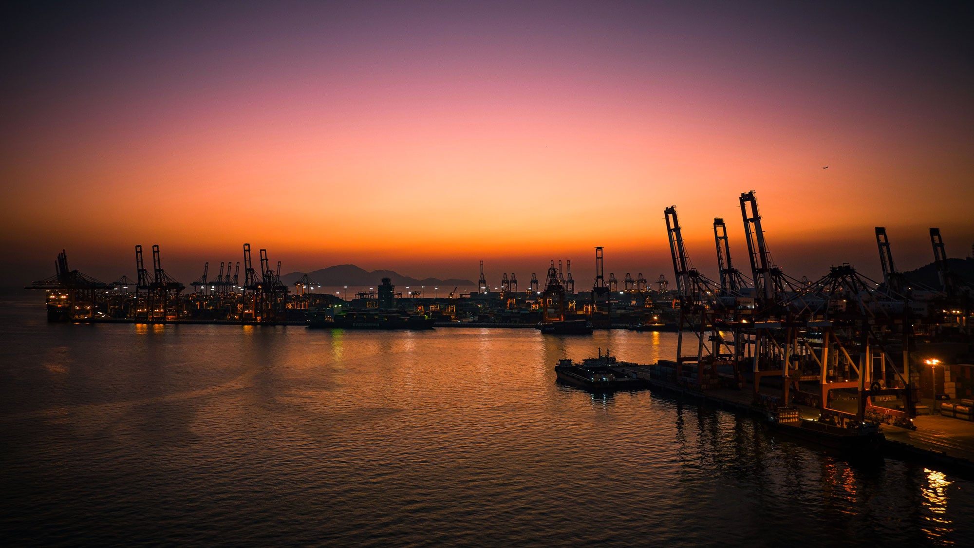 Aerial view of a busy shipping container port at sunset with cranes silhouetted against orange sky